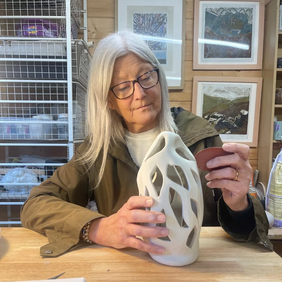 A woman with long grey hair and glasses holds a white pierced ceramic vase at a workbench