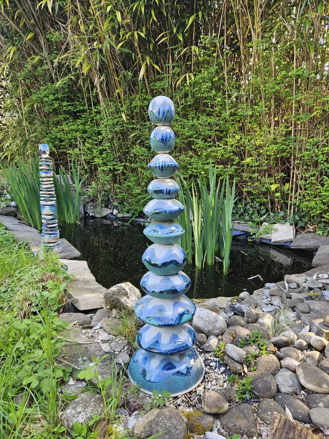 A tall stacked ceramic sculpture in blue glazes beside a garden pond