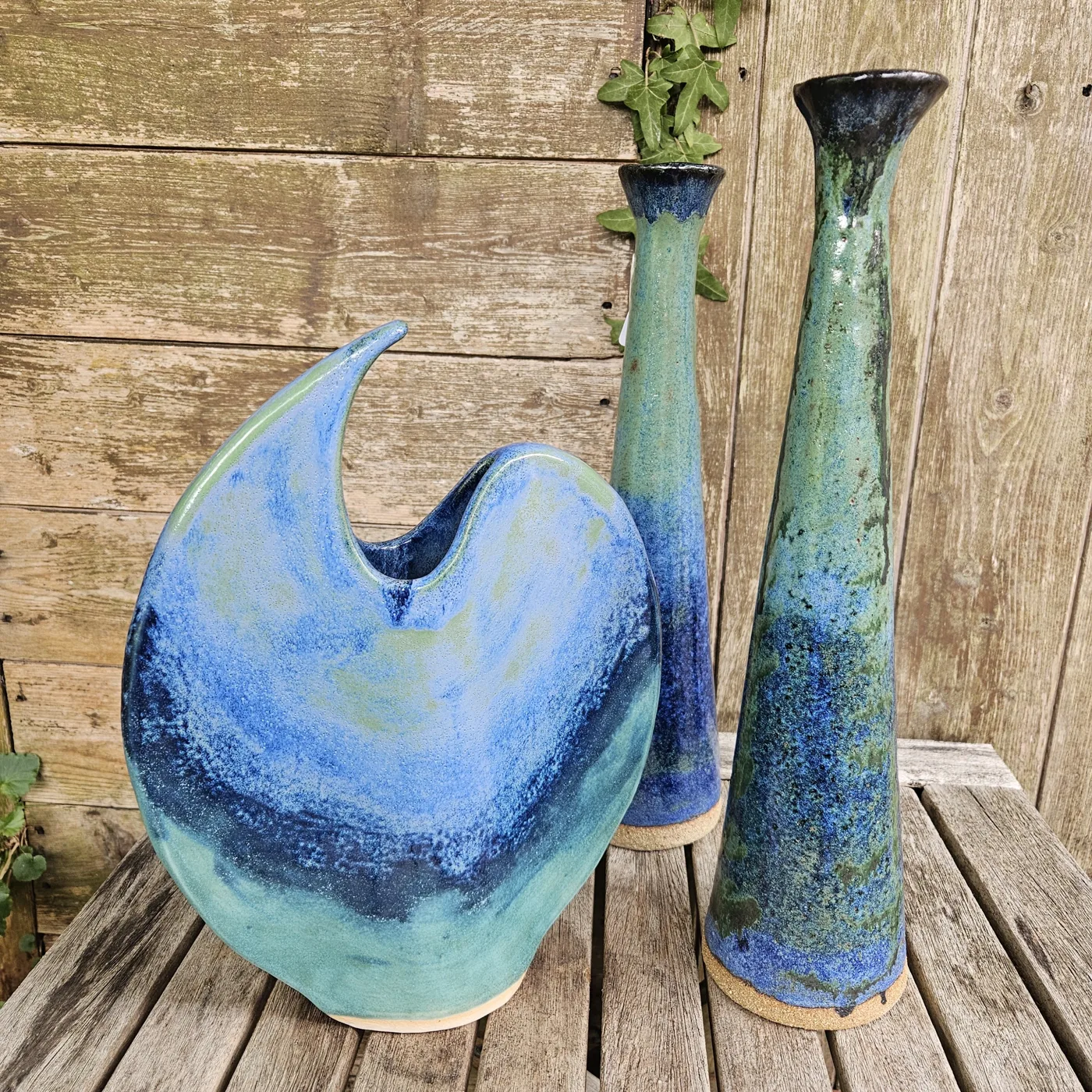 Three blue and green glazed ceramic vessels, one curved and two tall bottles, on a wooden table