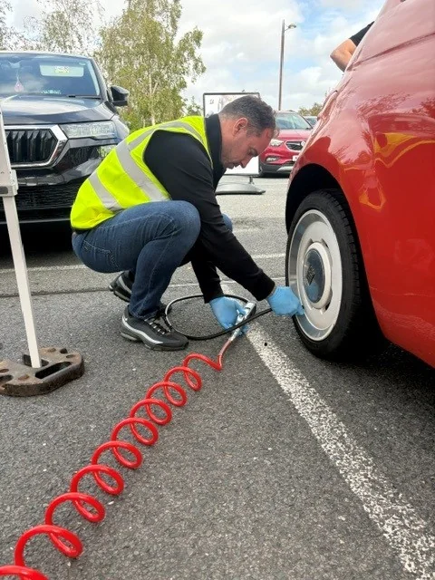 A technician in a high visibility vest crouching to check the tyre of a red car in a car park