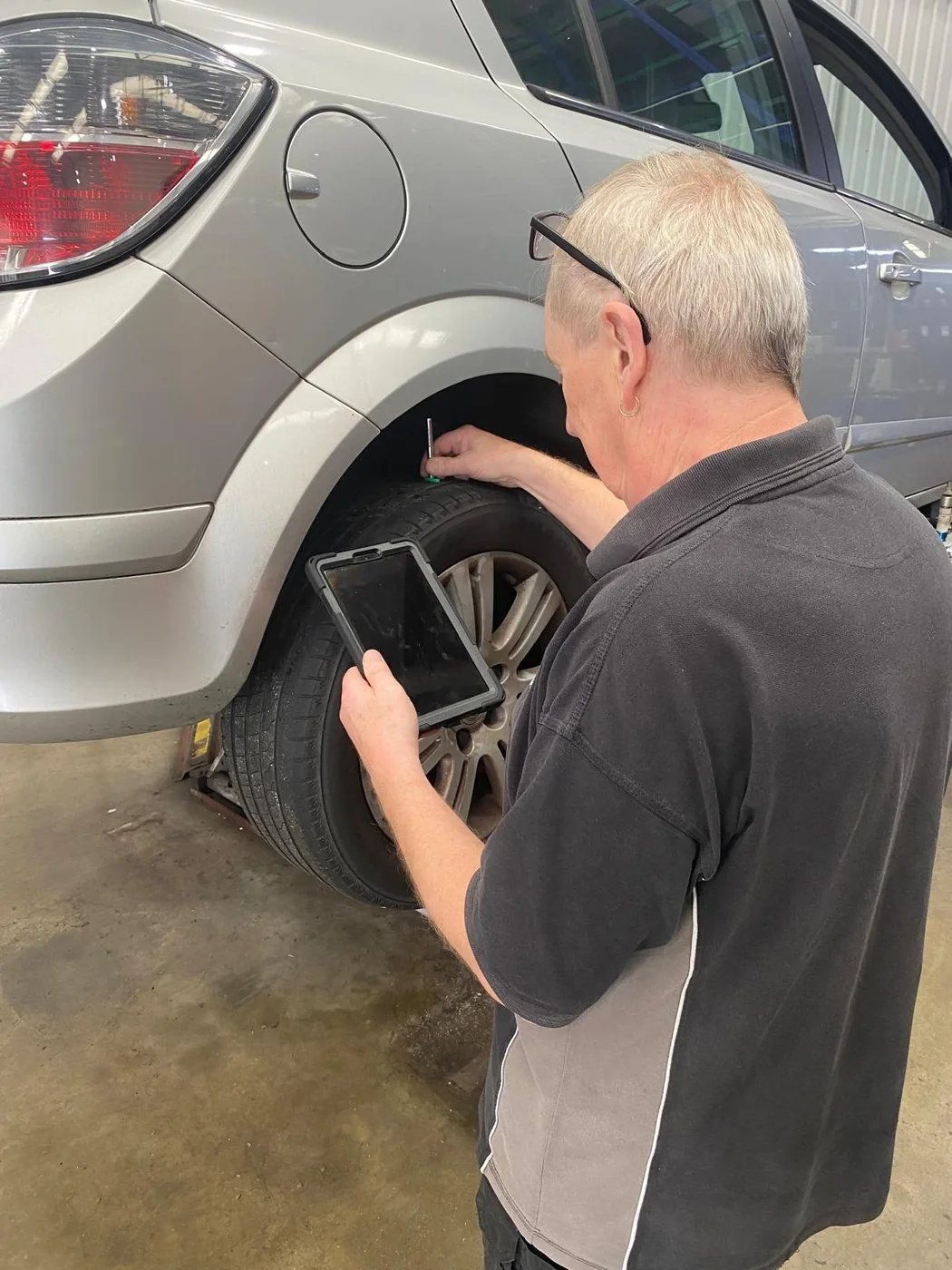 A technician holding a tablet while using a tread depth probe on the rear tyre of a silver car in a workshop